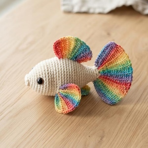 May include: A crocheted fish toy featuring a cream-coloured body with a dark eye and vibrant rainbow-coloured fins and tail. The fins and tail are worked in a fan-like pattern with distinct stripes of red, orange, yellow, green, blue, and purple yarn.