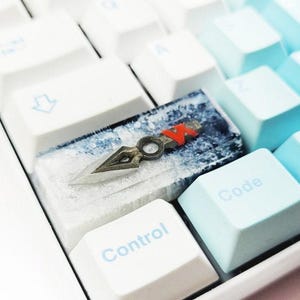 May include: A custom keyboard keycap featuring a small, decorative dagger with a red accent. The keycap is set on a white keyboard with light blue and white keys. The word "Control" is visible on a white key.