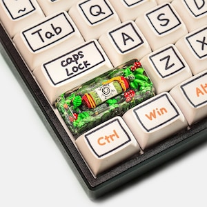 May include: Close-up of a keyboard with off-white keycaps. The "caps Lock" key is replaced with a custom keycap featuring a miniature forest scene with a decorative scroll. Other keys include "Tab", "Ctrl", "Win", and letter keys.