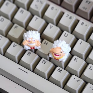 以下が含まれることがあります： ベージュ色のメカニカルキーボードのクローズアップ。2つのカスタムキーキャップが付いています。キーキャップは白で、笑顔の顔と巻き毛が特徴の漫画のキャラクターデザインです。キーボードのキーは標準的なQWERTYレイアウトで配置されています。
