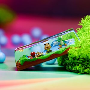 Puede incluir: Un terrario rectangular de vidrio con figuras en miniatura de personajes de videojuegos. La escena incluye un Yoshi verde, un Goomba marrón y un hongo rojo, sobre un fondo azul y blanco. El terrario está lleno de un líquido transparente.