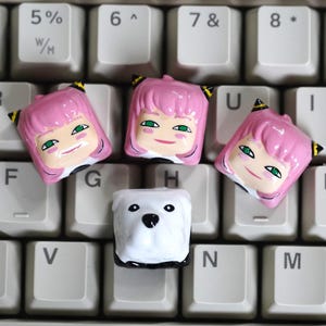 May include: A computer keyboard with custom keycaps. Three pink-haired character keycaps with green eyes and a smiling expression are arranged around a white bear keycap. The keyboard keys are white.