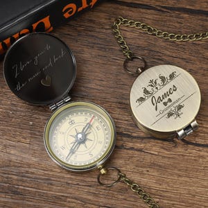 May include: Two antique-style compasses with chains on a wooden surface. One compass is open, showing a directional dial and the inscription "I love you to the moon and back." The other is closed and engraved with the name "James - Groomsman."