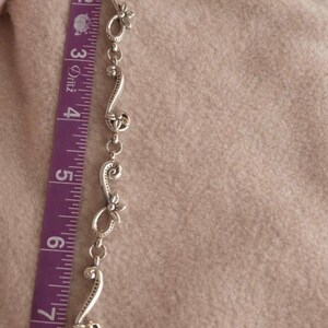 May include: A silver-toned bracelet with floral and scroll design elements, approximately 7 inches long. The bracelet is measured with a purple and white measuring tape. The background is a soft, neutral color.