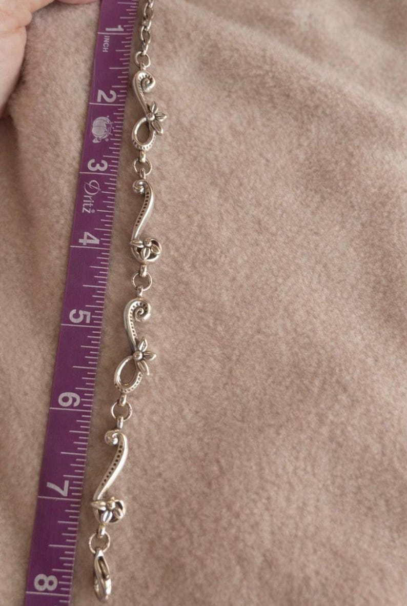 May include: A silver-toned bracelet with floral and scroll design elements, approximately 7 inches long. The bracelet is measured with a purple and white measuring tape. The background is a soft, neutral color.