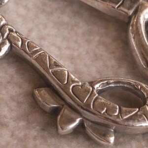 May include: Close-up of a silver-toned metal pendant with a key design. The key features heart-shaped cutouts and floral detailing. The pendant is on a neutral-toned surface, highlighting the intricate design and metallic sheen.