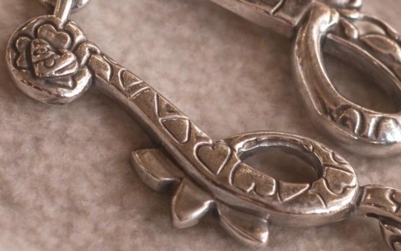 May include: Close-up of a silver-toned metal pendant with a key design. The key features heart-shaped cutouts and floral detailing. The pendant is on a neutral-toned surface, highlighting the intricate design and metallic sheen.