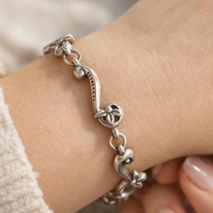 May include: A silver bracelet with a floral and scroll design. The bracelet features a series of decorative links, each with a flower or scroll motif. The bracelet is made of a shiny, metallic material and is worn on a wrist.