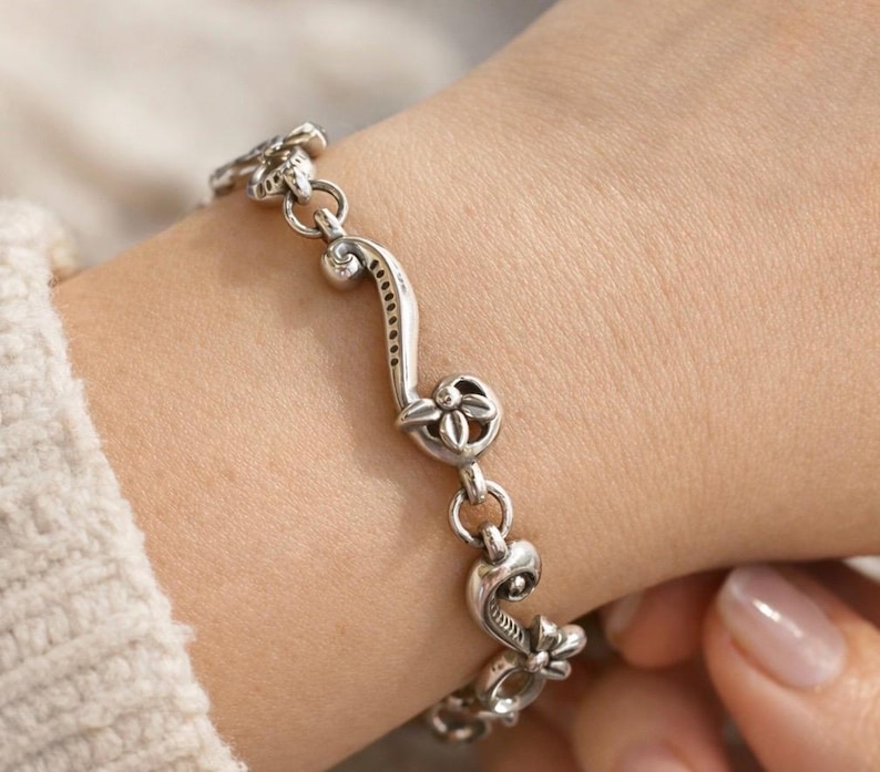 May include: A silver bracelet with a floral and scroll design. The bracelet features a series of decorative links, each with a flower or scroll motif. The bracelet is made of a shiny, metallic material and is worn on a wrist.