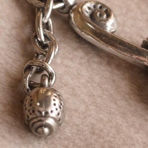 May include: Close-up of a silver-toned metal charm with a decorative design. The charm features a chain, a small, perforated acorn-shaped pendant, and a stylized, curved element with a scroll design. The background is a soft, neutral color.