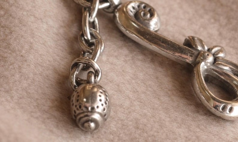 May include: Close-up of a silver-toned metal charm with a decorative design. The charm features a chain, a small, perforated acorn-shaped pendant, and a stylized, curved element with a scroll design. The background is a soft, neutral color.