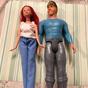 May include: Two dolls are displayed. One doll has red hair, a white top with a red emblem, and blue pants. The other doll has blonde hair, a blue shirt, and gray pants. Both dolls are plastic and have articulated limbs.