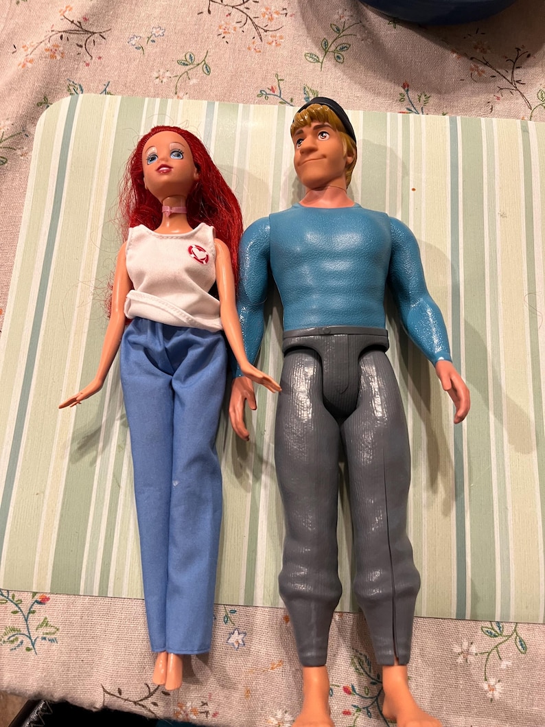 May include: Two dolls are displayed. One doll has red hair, a white top with a red emblem, and blue pants. The other doll has blonde hair, a blue shirt, and gray pants. Both dolls are plastic and have articulated limbs.