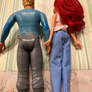 May include: Two vintage dolls are shown from the back. One doll has blonde hair, a blue top, gray pants, and a black cap. The other doll has red hair, a white top, and blue pants. The dolls are standing on a striped surface.