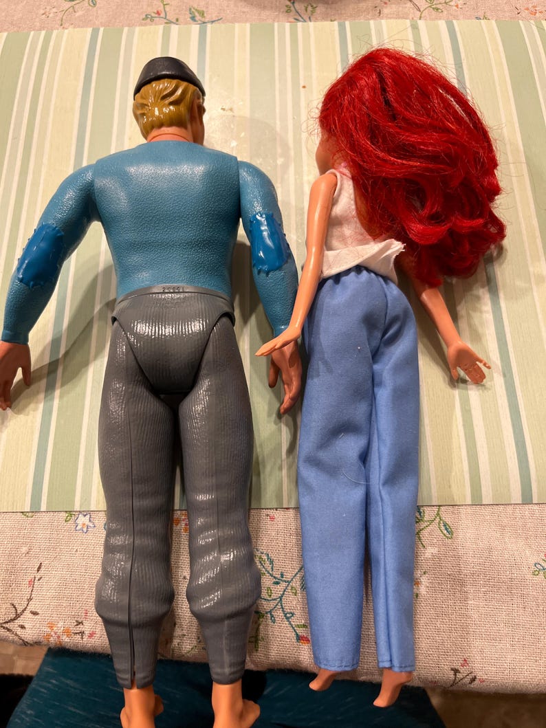 May include: Two vintage dolls are shown from the back. One doll has blonde hair, a blue top, gray pants, and a black cap. The other doll has red hair, a white top, and blue pants. The dolls are standing on a striped surface.