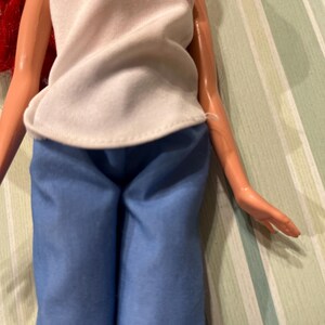 May include: A doll wearing a white sleeveless top with a pink heart design and blue pants. The doll has red hair and is posed against a striped background.