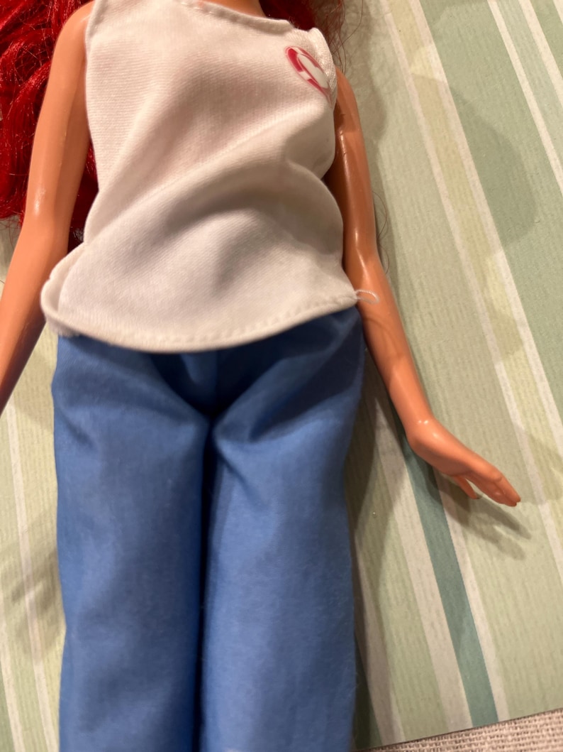May include: A doll wearing a white sleeveless top with a pink heart design and blue pants. The doll has red hair and is posed against a striped background.