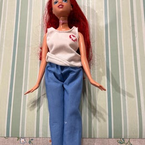 May include: A doll with red hair, blue eyes, and a pink choker. The doll is wearing a white tank top with a red heart design and blue pants. The doll is standing in front of a striped background.