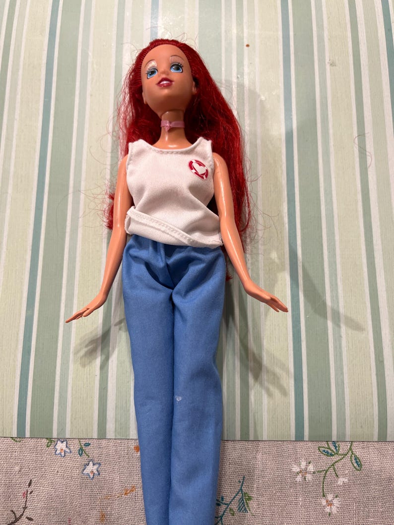 May include: A doll with red hair, blue eyes, and a pink choker. The doll is wearing a white tank top with a red heart design and blue pants. The doll is standing in front of a striped background.