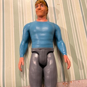 May include: A vintage action figure of a man with blonde hair, wearing a blue long-sleeve shirt, gray pants, and a black cap. The figure has a neutral expression and is standing upright. The figure is made of plastic.