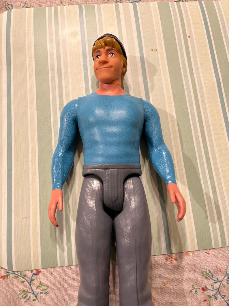 May include: A vintage action figure of a man with blonde hair, wearing a blue long-sleeve shirt, gray pants, and a black cap. The figure has a neutral expression and is standing upright. The figure is made of plastic.