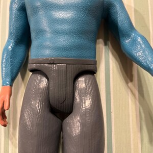 May include: A vintage action figure featuring a blue textured top and gray pants. The figure has orange skin-toned hands and a neutral expression. The toy is a collectible item.