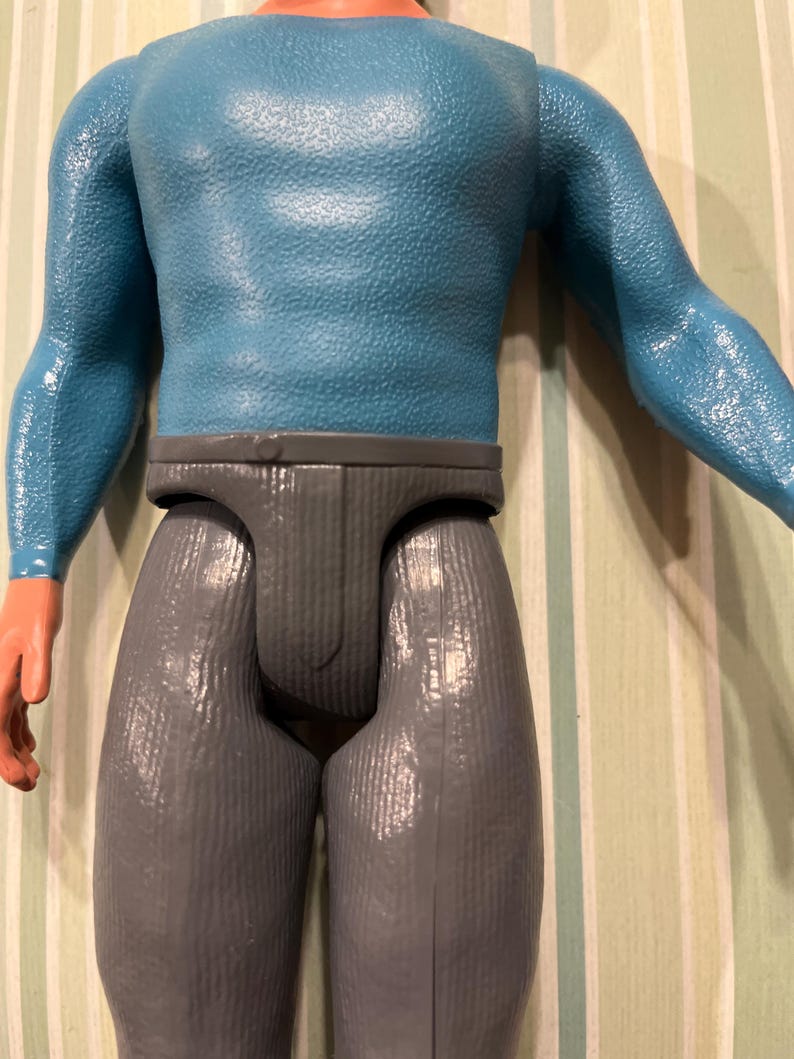 May include: A vintage action figure featuring a blue textured top and gray pants. The figure has orange skin-toned hands and a neutral expression. The toy is a collectible item.