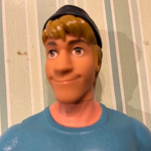 May include: A plastic figurine of a man with blonde hair, blue eyes, and a slight smile. He wears a black cap and a light blue shirt. The figurine is a close-up shot.