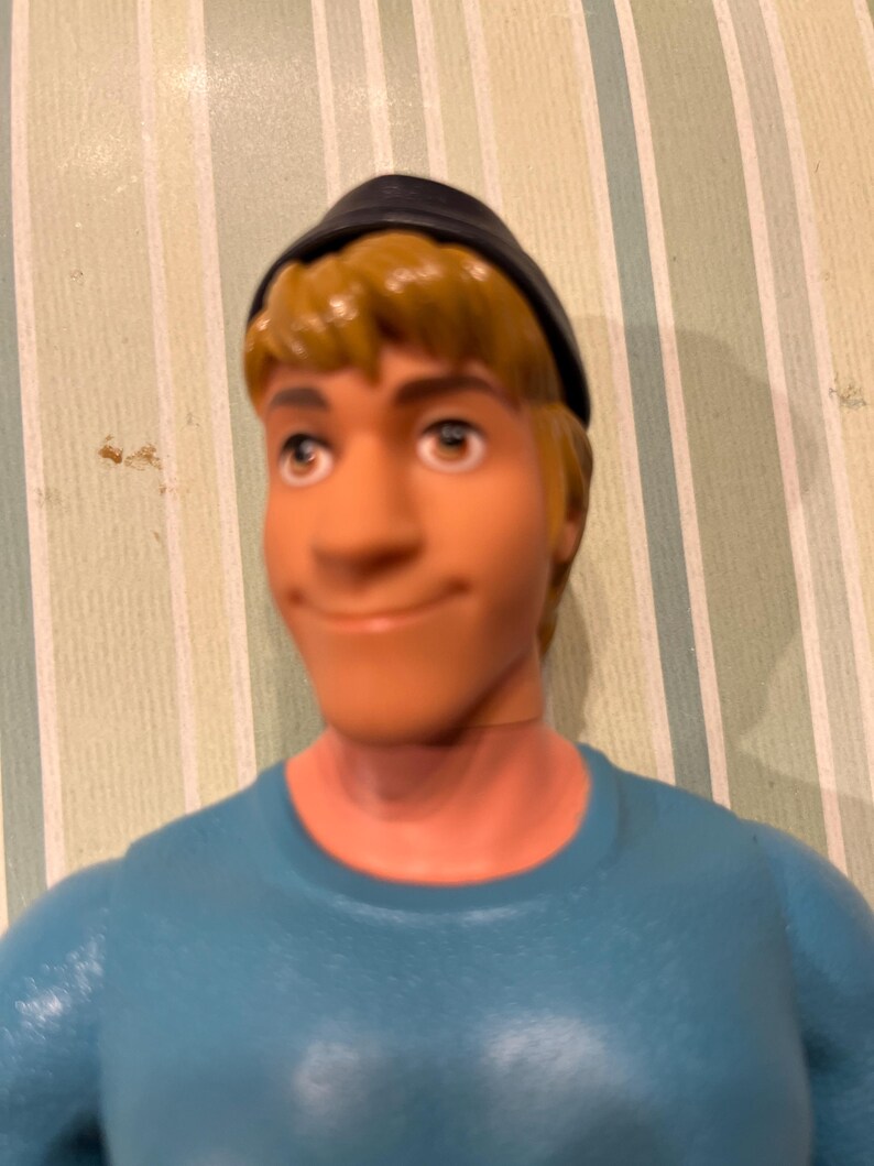 May include: A plastic figurine of a man with blonde hair, blue eyes, and a slight smile. He wears a black cap and a light blue shirt. The figurine is a close-up shot.
