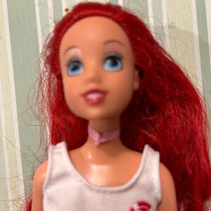 May include: A doll with bright red hair, blue eyes, and a pink choker. The doll wears a white top with a red and white heart-shaped emblem. The doll's face has a cheerful expression.