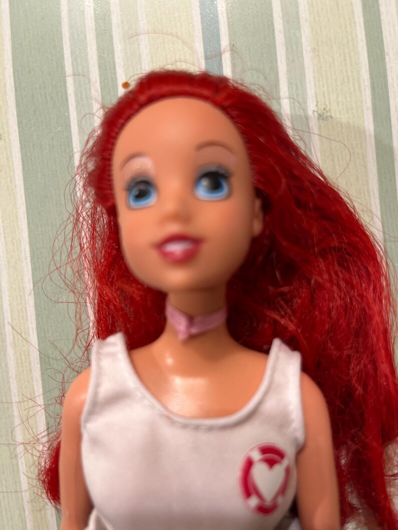 May include: A doll with bright red hair, blue eyes, and a pink choker. The doll wears a white top with a red and white heart-shaped emblem. The doll's face has a cheerful expression.