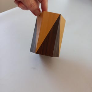 Peut inclure: Un bloc de bois géométrique avec des panneaux en bois brun clair et foncé. Le bloc est tenu par une main, mettant en valeur son design angulaire et ses couleurs contrastées. L'objet semble être un objet décoratif ou une petite sculpture.