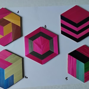 Puede incluir: Cinco posavasos hexagonales con diseños geométricos. Los posavasos presentan colores rosa, negro, verde, azul y amarillo. Cada posavasos tiene un patrón único. Se muestran sobre una superficie blanca.