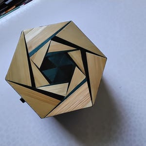 Op de afbeelding: Een zeshoekige houten doos met een uniek ontwerp. De doos heeft lichtbruine houten panelen in een spiraalpatroon, met donkerblauwe accenten en zwarte lijnen. Waarschijnlijk gebruikt voor opslag of presentatie, met een geometrische uitstraling.