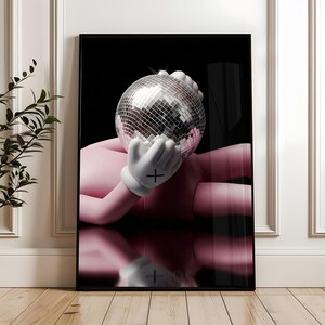 May include: A framed art print featuring a pink figure holding a silver disco ball. The figure wears white gloves with a black cross on the palm. The background is black, and the image is reflected below.