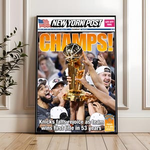 May include: A framed New York Post newspaper front page featuring the headline CHAMPS! in large yellow letters. The image shows basketball players holding a gold trophy aloft, with the text Knicks fans rejoice as team wins first title in 53 years.
