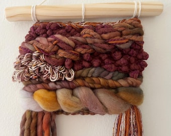 May include: A woven wall hanging with a wooden dowel. The textile art piece features a variety of textures and colors, including burgundy, orange, yellow, and cream. The piece is made of thick yarn and fiber, creating a tactile and visually interesting design.