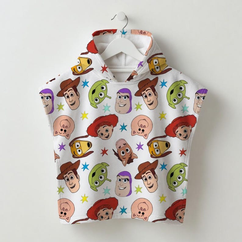 May include: White hooded poncho featuring a repeating pattern of cartoon character faces and colorful stars. The characters include a cowboy, a space ranger, and other familiar faces. The poncho is hanging on a white hanger.