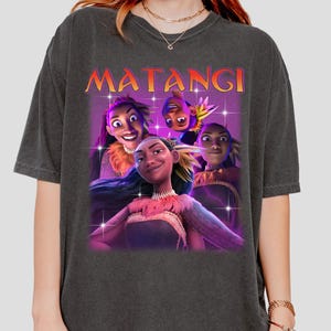 Op de afbeelding: Donkergrijs T-shirt met een afbeelding van vier geanimeerde personages en het woord "MATANGI" in oranje. De personages hebben verschillende huidtinten en dragen decoratieve kleding. De afbeelding heeft een paarse en roze achtergrond met stervormige glitters.