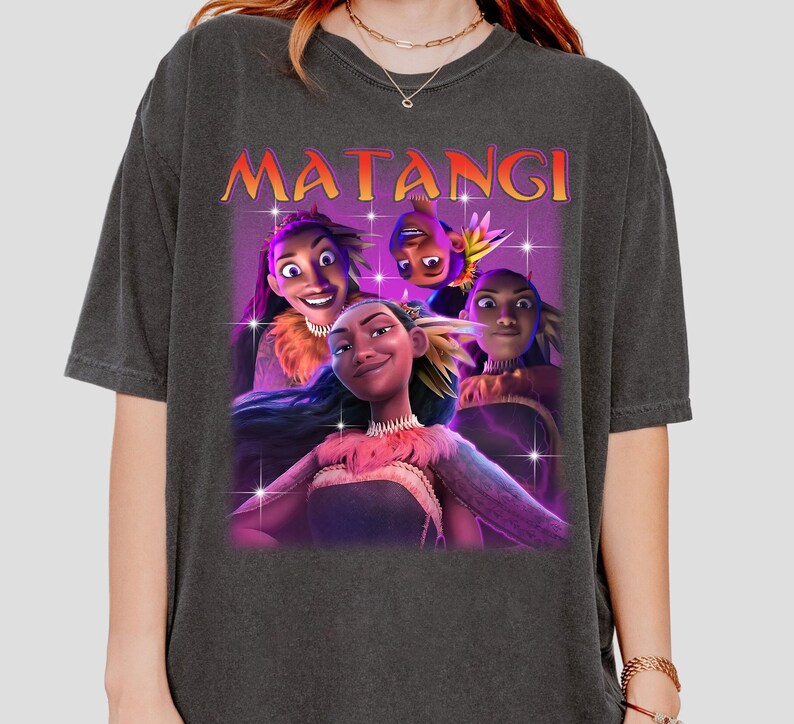 Op de afbeelding: Donkergrijs T-shirt met een afbeelding van vier geanimeerde personages en het woord "MATANGI" in oranje. De personages hebben verschillende huidtinten en dragen decoratieve kleding. De afbeelding heeft een paarse en roze achtergrond met stervormige glitters.