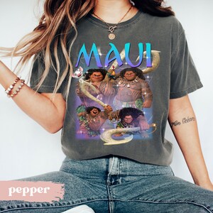 May include: Dark gray t-shirt featuring the word "MAUI" in teal and purple, with a graphic of the character Maui from the animated film. The design includes images of Maui holding his magical fish hook. A pig is also featured in the design.