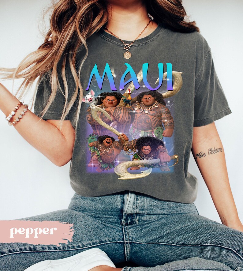 May include: Dark gray t-shirt featuring the word "MAUI" in teal and purple, with a graphic of the character Maui from the animated film. The design includes images of Maui holding his magical fish hook. A pig is also featured in the design.