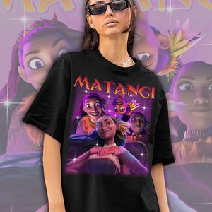 Op de afbeelding: Zwart T-shirt met een afbeelding van animatiefiguren en het woord "MATANGI" in oranje. Het ontwerp heeft een paarse en roze achtergrond met sprankelende effecten. Het shirt wordt gedragen door een persoon met een zonnebril.
