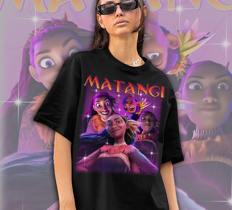 Op de afbeelding: Zwart T-shirt met een afbeelding van animatiefiguren en het woord "MATANGI" in oranje. Het ontwerp heeft een paarse en roze achtergrond met sprankelende effecten. Het shirt wordt gedragen door een persoon met een zonnebril.
