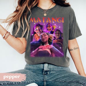 Op de afbeelding: Een grijze t-shirt met een afbeelding van geanimeerde personages en het woord "MATANGI" in oranje en geel. De personages staan tegen een paarse achtergrond met sterachtige accenten. Het t-shirt wordt door een persoon gedragen.