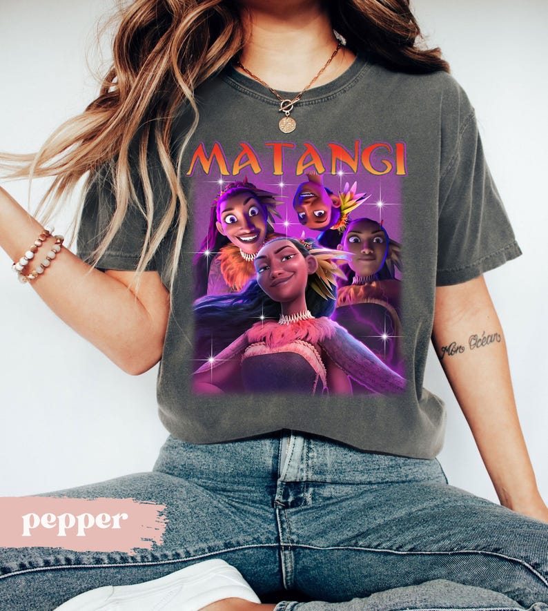 Op de afbeelding: Een grijze t-shirt met een afbeelding van geanimeerde personages en het woord "MATANGI" in oranje en geel. De personages staan tegen een paarse achtergrond met sterachtige accenten. Het t-shirt wordt door een persoon gedragen.