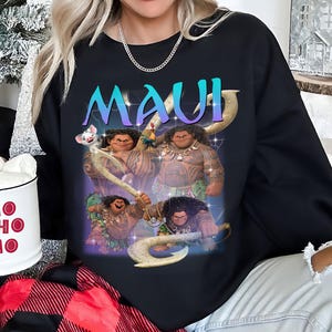 May include: Black sweatshirt featuring a graphic of the character Maui from the animated film. The word "MAUI" is displayed in a gradient blue and purple font. The design includes images of Maui in various poses with his signature hook.