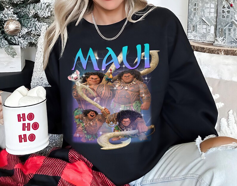 May include: Black sweatshirt featuring a graphic of the character Maui from the animated film. The word "MAUI" is displayed in a gradient blue and purple font. The design includes images of Maui in various poses with his signature hook.