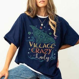 May include: Navy blue t-shirt with the text "Village Crazy Lady" in a colorful design. The shirt features a stylized manta ray graphic in teal and green, with floral accents. The shirt is a casual, comfortable style.