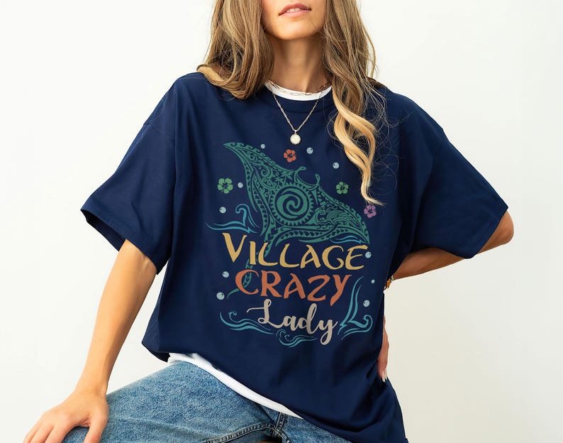 May include: Navy blue t-shirt with the text "Village Crazy Lady" in a colorful design. The shirt features a stylized manta ray graphic in teal and green, with floral accents. The shirt is a casual, comfortable style.